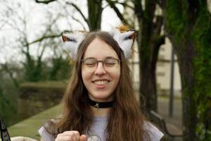 A photo showing Anna Aurora outdoors from the top to her sholders. She is wearing fox ears, a collar and a grey tshirt. She is holding her right hand up to her shoulder in joy. She has her eyes closed and is smiling. The background contains the sky, mossy